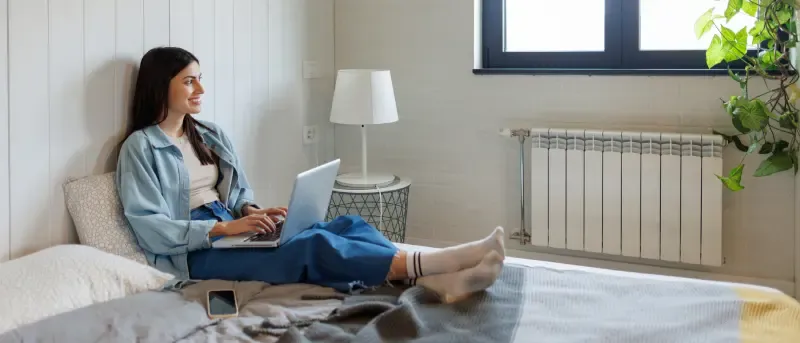 A smiling young woman sits comfortably on a bed with a laptop on her knees, working or browsing.